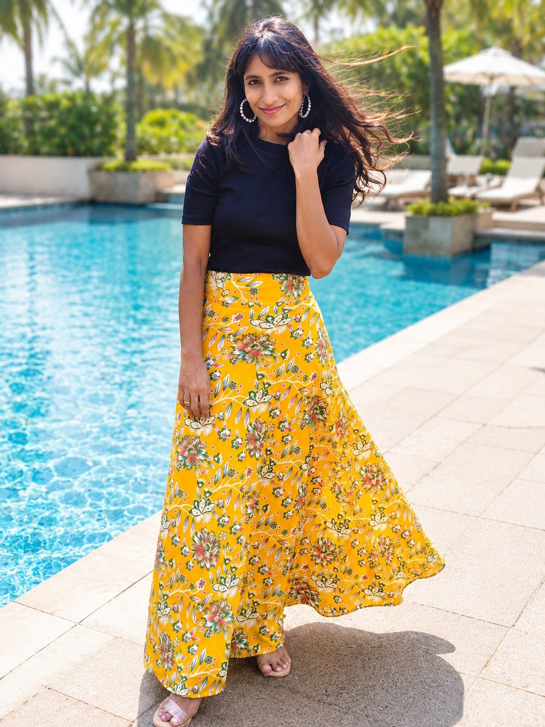 Woman in a yellow floral skirt and black top standing by a poolside.