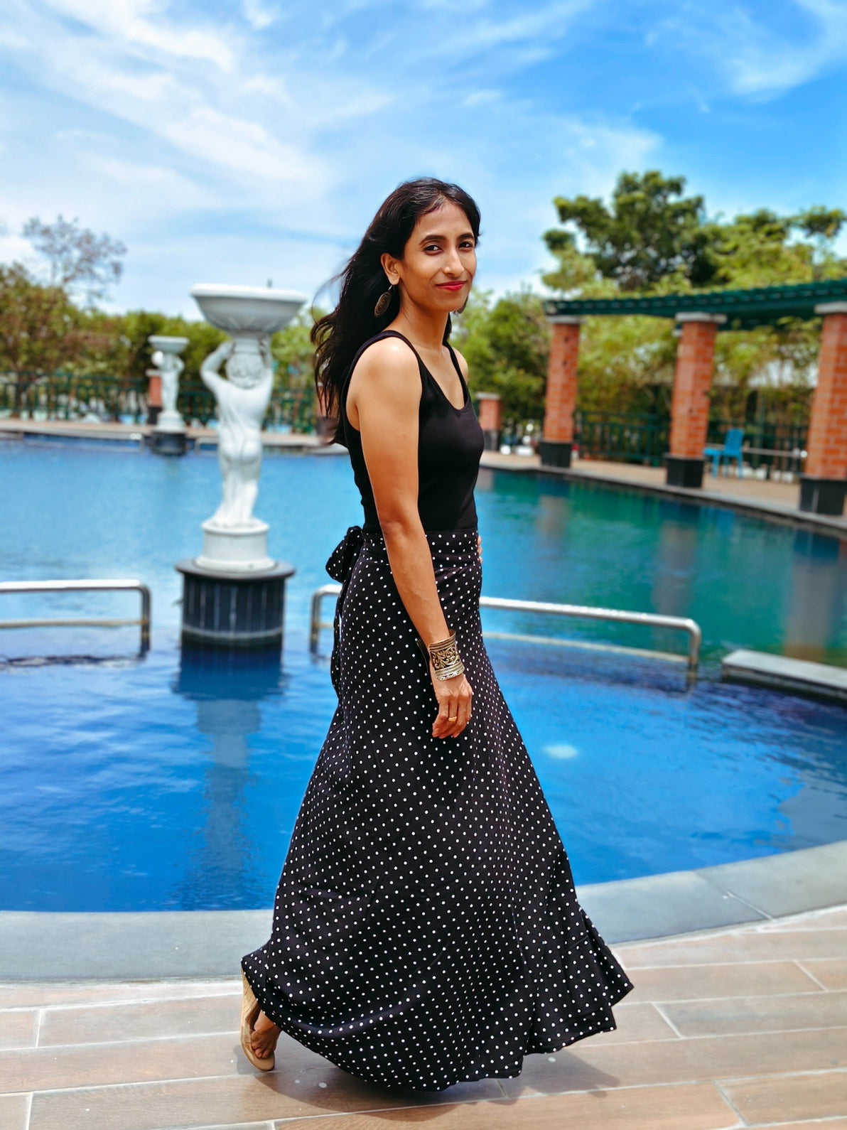 Woman in a black polka dot skirt standing by a pool with trees and a clear sky in the background